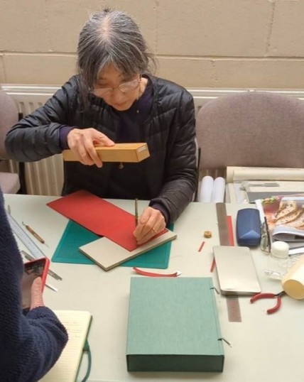 Midori Kunikata-Cockram using a rectangle piece of wood to hit a chisel creating a slot in the red covered case laid flat.

The example box covered in light green cloth sits at the front of the table.