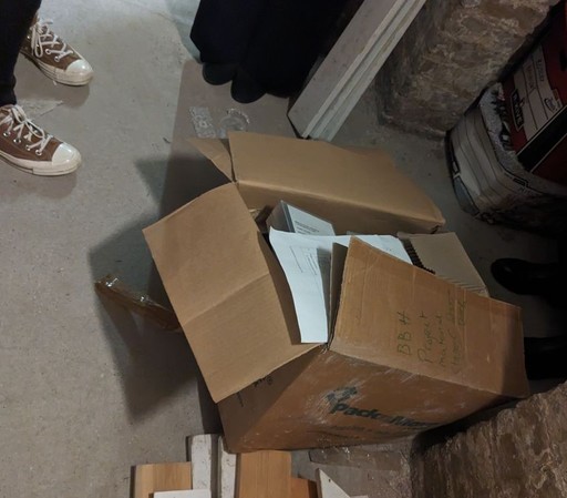 A brown cardboard box with separating seams sitting on a cellar floor amongst building detritus.