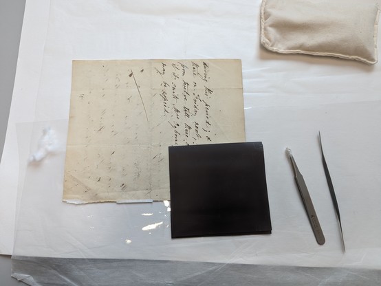 A A5ish size cream paper flat on a surface lined with blotter. Brown/black manuscript across half the page. A brown square located at the right lower corner. A pair of tweezers and micro-spatula arranged to the right.