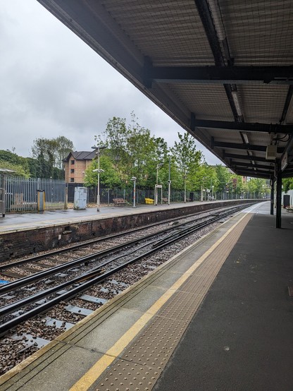 Typical urban train station: asphalt platform surface, yellow standing line, multiple types of fencing including chain link, rain and grey.
