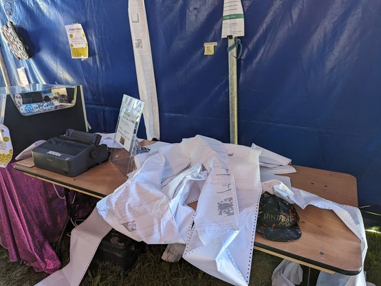 Blue festival tent.
Wood table.
Black dot matrix printer sitting on left of table.
Remainder of table covered in lengths of paper.