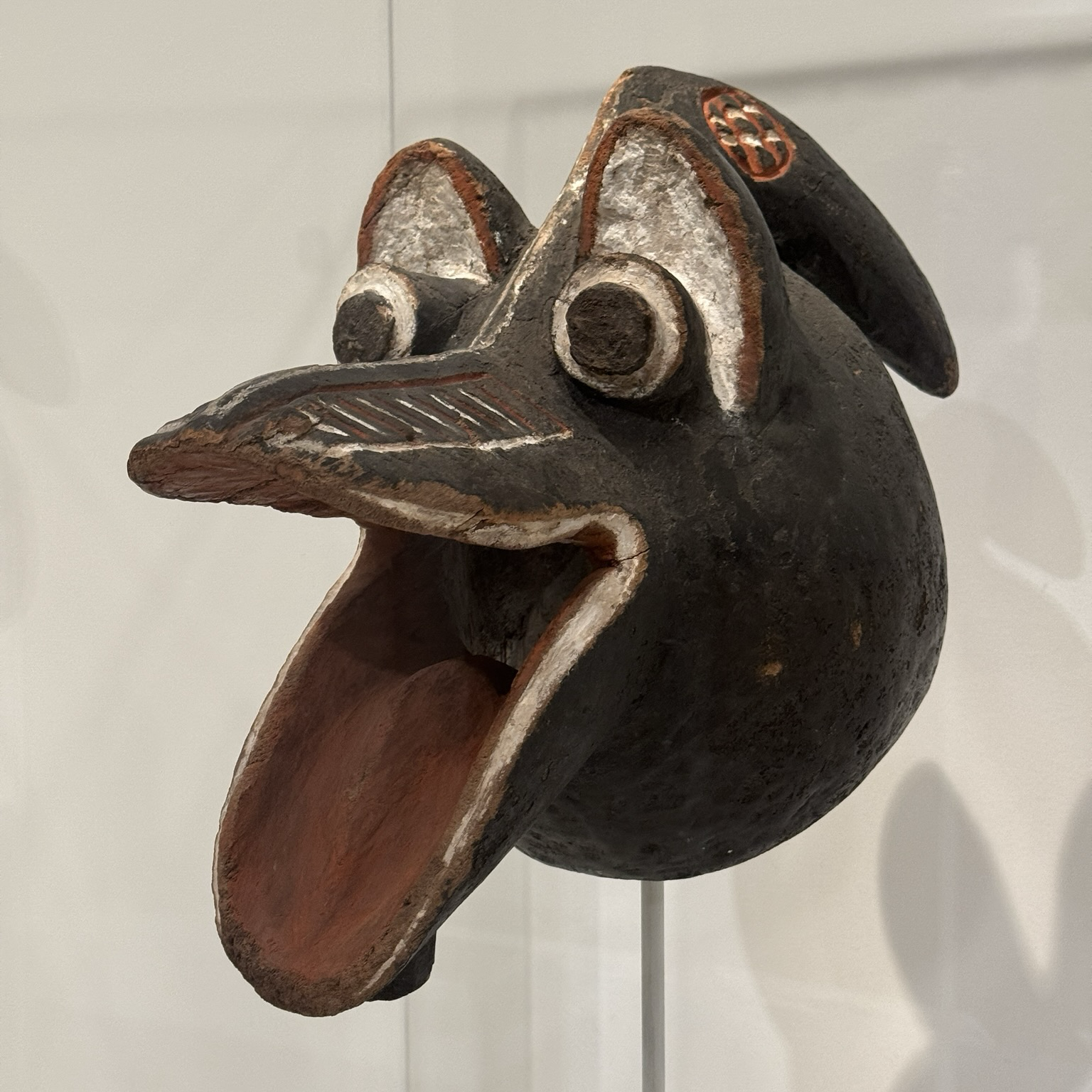 photo of the crow mask mounted on display at museum, quarter turn side profile view painted black with red and white details happy looking crow head! big googly eyes and wide open mouth like :D