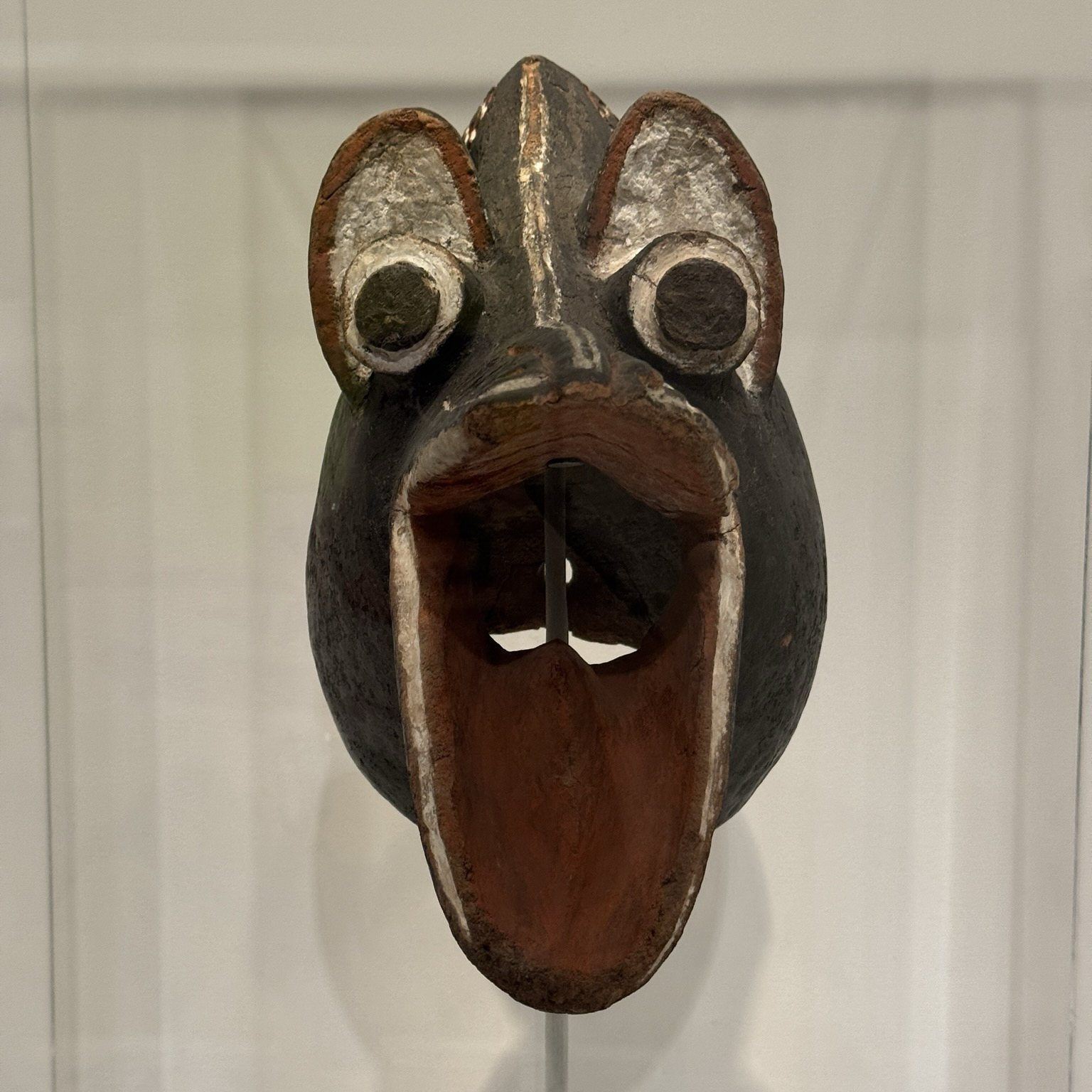 photo of the crow mask mounted on display at museum, front profile view painted black with red and white details happy looking crow head! big googly eyes and wide open mouth like :D