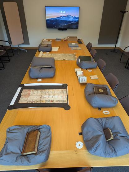 Leather bound volumes, manuscripts, positioned on grey cushions along a long wooden table. Several large scale facsimiles interspersed between original collection items.